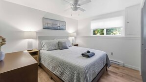 Un dormitorio con cama, ventilador de techo, mesita de noche y una ventana con persianas, además de un cuadro en la pared.