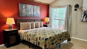A bedroom with a bed, two bedside tables with lamps, a window with curtains, a wall-mounted fan, and a painting.