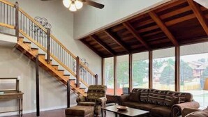 A spacious living room with a wooden staircase, a large window, and a ceiling fan.