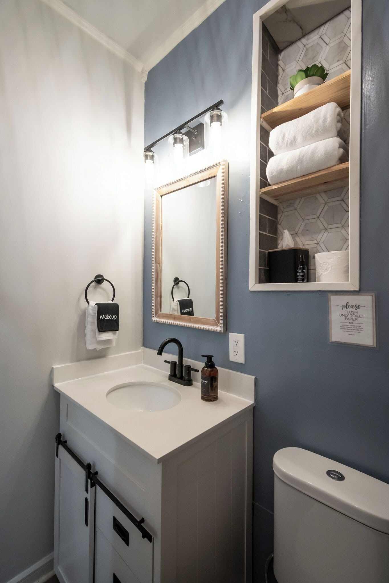 Une salle de bain avec un lavabo blanc, un robinet noir et un miroir doté d’un cadre décoratif. On y trouve un porte-serviettes avec une serviette étiquetée « Maquillage » et un panneau indiquant : « Veuillez jeter le papier toilette dans les toilettes ».