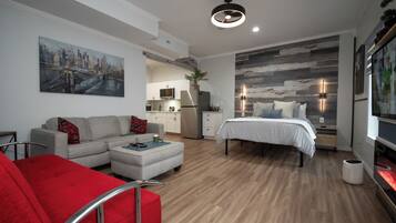 Una sala moderna con sofá, un sillón rojo, una cama y una cocina al fondo.