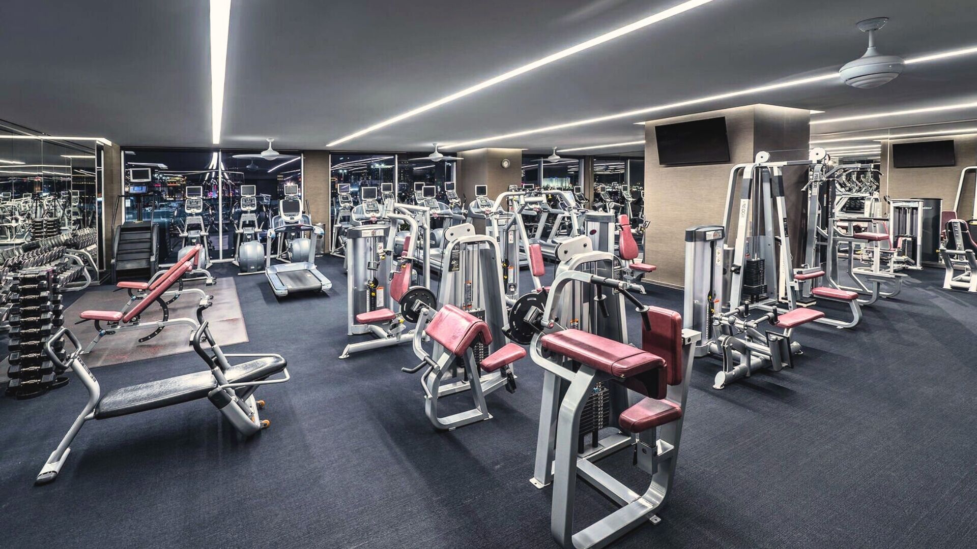 A modern gym with various exercise machines, mirrors, and a dark carpeted floor.