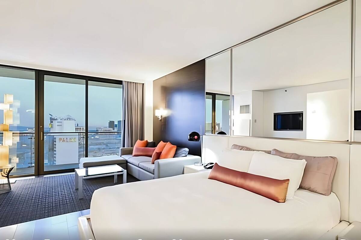 A modern hotel room with a large bed, a sofa, a TV, and a city view.