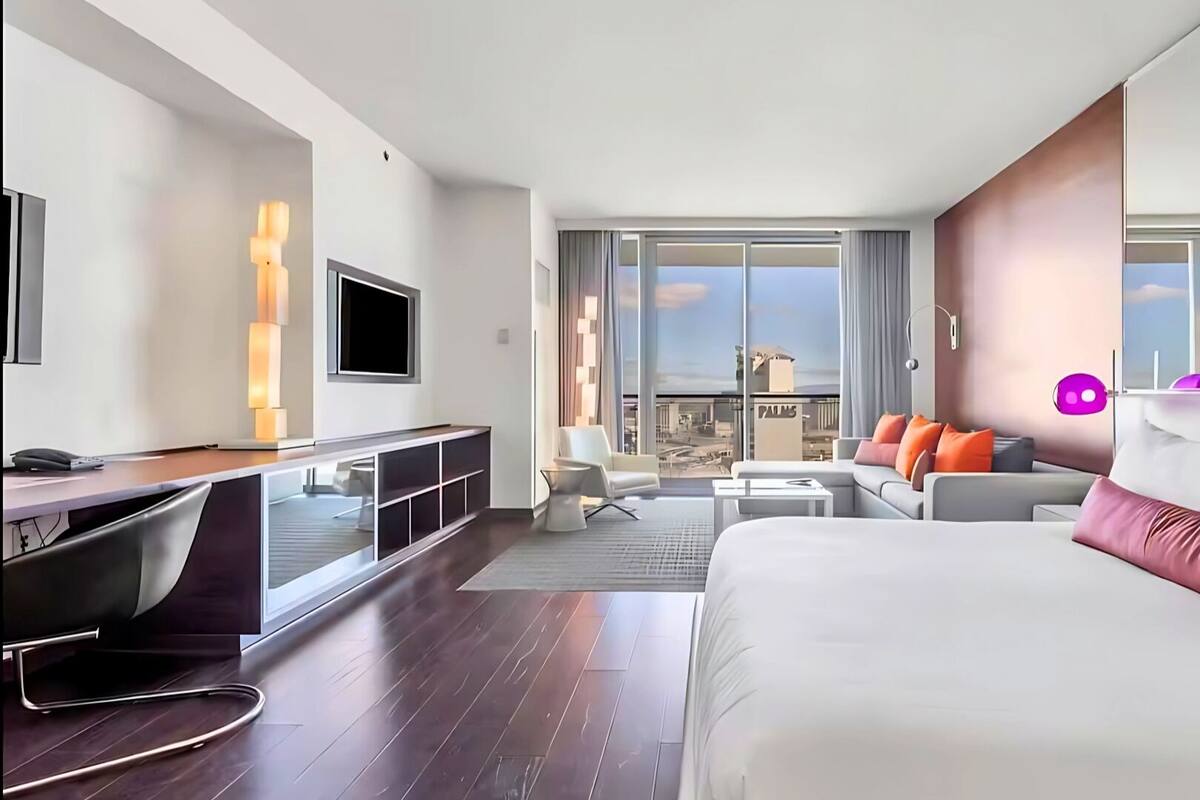 A modern hotel room with a large bed, a desk, a sofa, and a balcony with a city view.