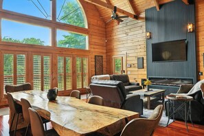 A spacious living room with a large wooden dining table, comfortable seating, a fireplace, and a television mounted on the wall. - Secluded Nashville Luxury Lodge (Ashland City)