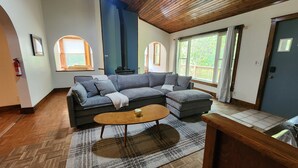 Ein Wohnzimmer mit einem Ecksofa, einem Couchtisch und großen Fenstern mit Vorhängen.