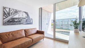Un salon moderne avec un canapé en cuir marron, une grande fenêtre donnant sur un paysage urbain et une photographie en noir et blanc d’un pont accrochée au mur.
