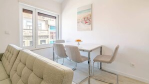 A modern living room with a sofa, a dining table with chairs, a window with a view of buildings, and a painting on the wall. - Charming Mezzanine Studio by HostWise (Porto)