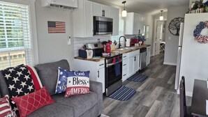 A modern kitchen with white cabinets, a microwave, and a stove. There is a gray sofa with American-themed pillows and a rug with a patriotic pattern. - Liberty Nook Tiny Home (Big Cabin)