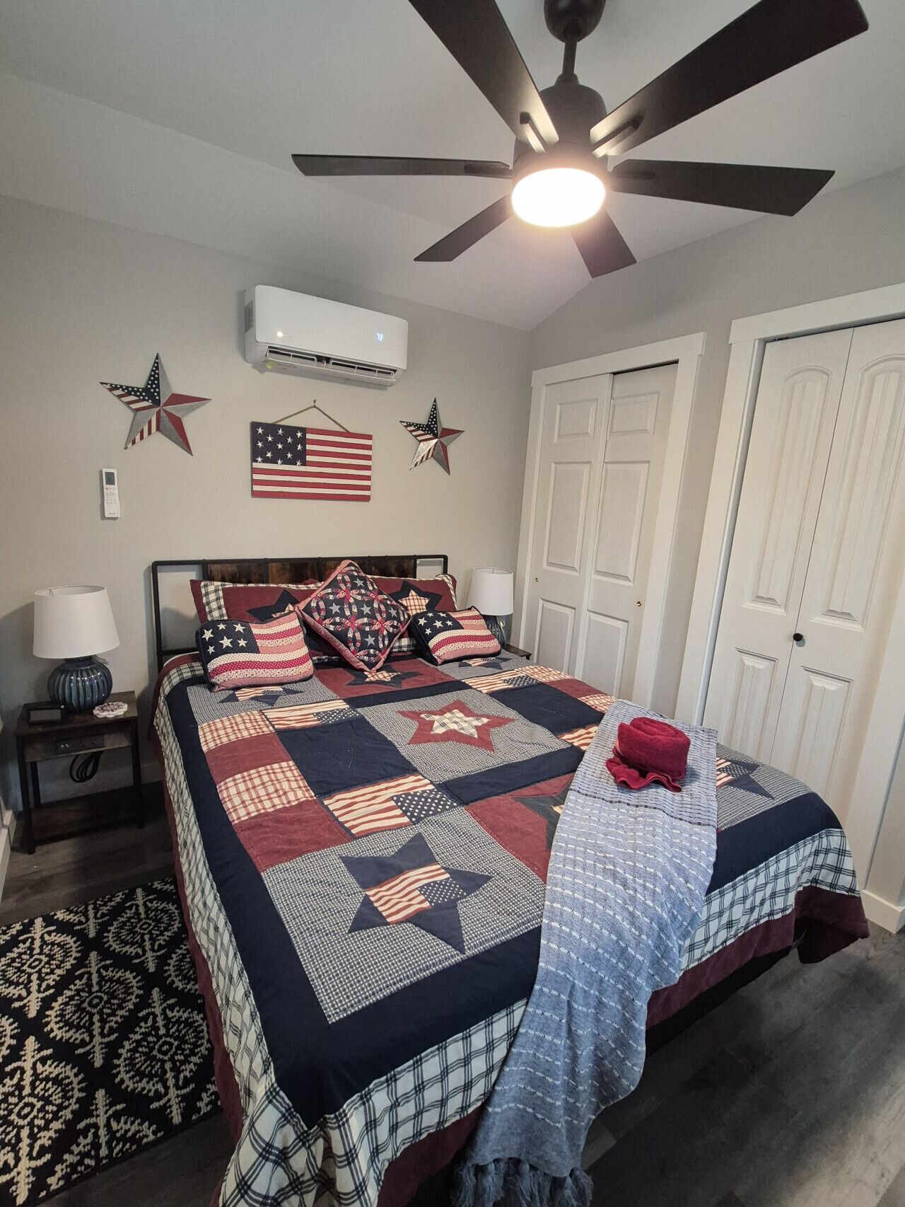 Une chambre à coucher comprenant un lit, un ventilateur de plafond, un appareil de climatisation, un drapeau décoratif et un tapis à motifs.