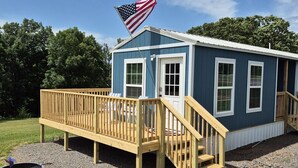 A small, single-story building with a wooden deck and a flagpole with an American flag. - Liberty Nook Tiny Home (Big Cabin)