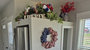 A kitchen with a stainless steel refrigerator, a white wall with a patriotic wreath, and a window with blinds. - Liberty Nook Tiny Home (Big Cabin)