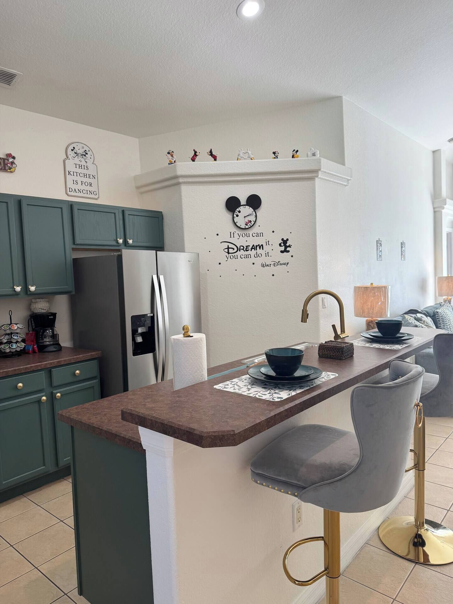 Une cuisine moderne avec des meubles de couleur verte, un réfrigérateur en acier inoxydable, un plan de travail en marbre et un autocollant mural sur le thème de Mickey Mouse.