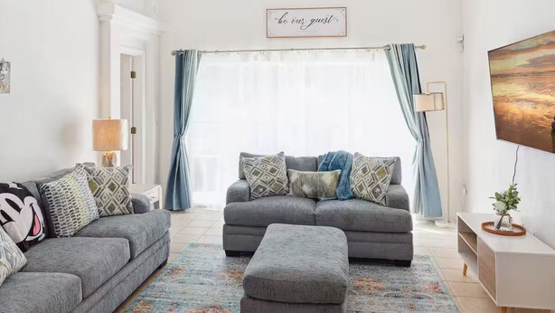 A living room with two grey sofas, a matching ottoman, a patterned rug, a painting, and a 'be our guest' sign.