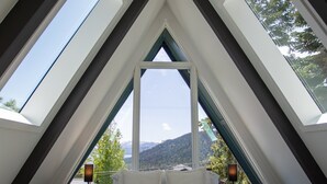A bedroom with a sloped ceiling and skylights, a bed, bedside tables, and a view of a mountainous landscape.