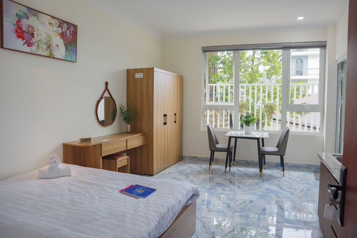 Habitación de hotel con cama, escritorio, silla, armario y ventana con vistas a un balcón.