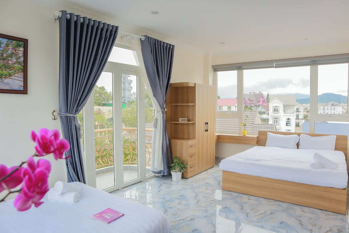 Habitación de hotel con cama, armario de madera, planta en maceta y vistas a la ciudad a través de amplios ventanales.