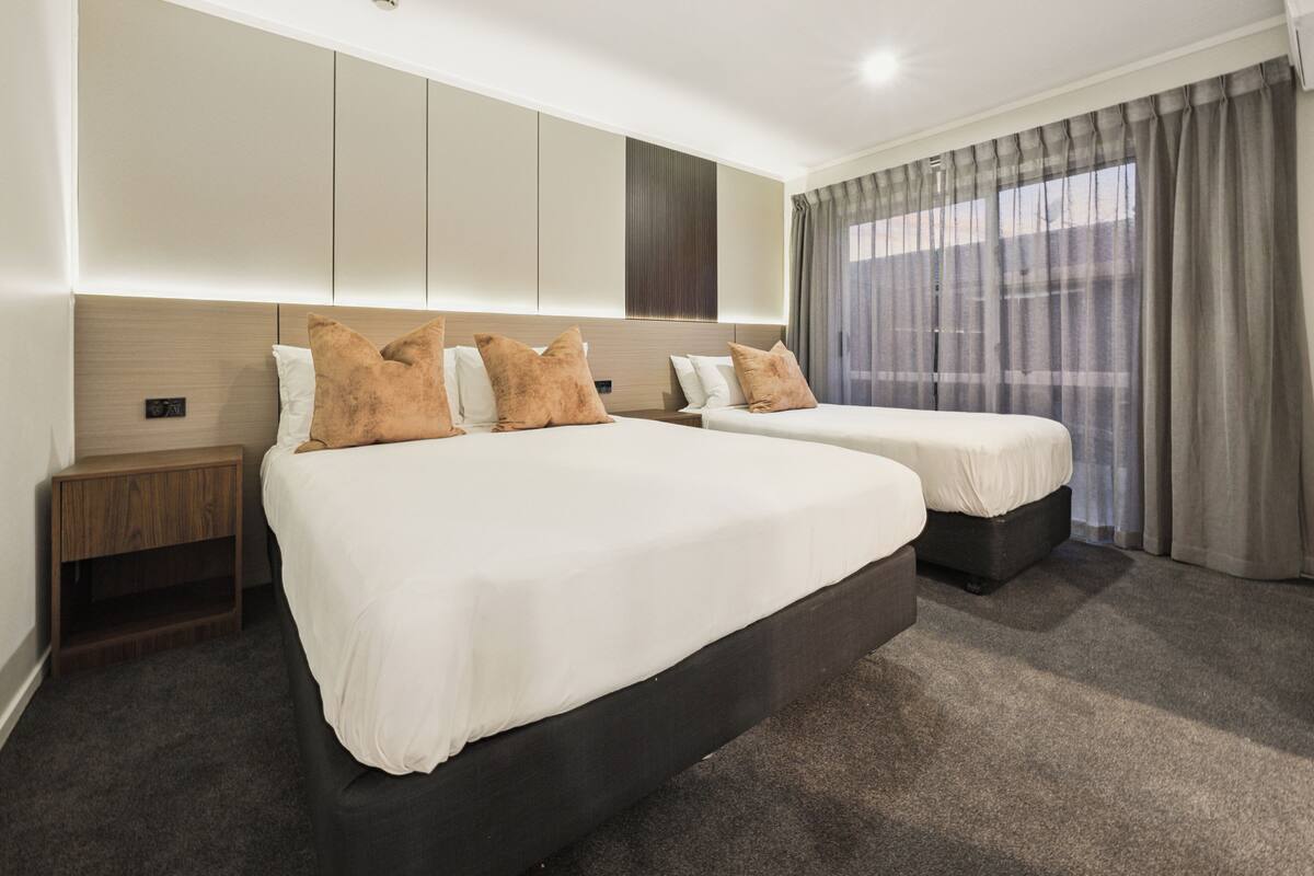 A hotel room with two beds, a wooden headboard, a small wooden nightstand, and a large window with curtains.