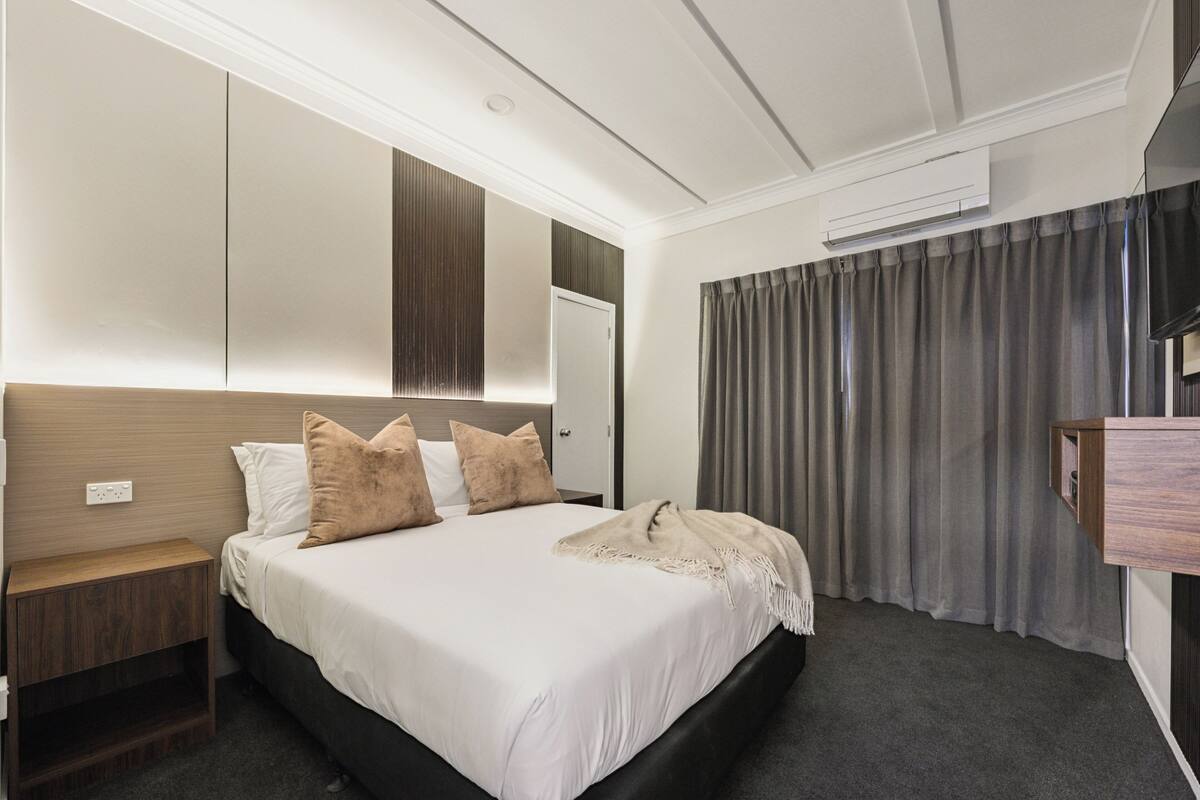 A hotel room with a large bed, a wooden nightstand, a mirror, a wardrobe, and a window with curtains.