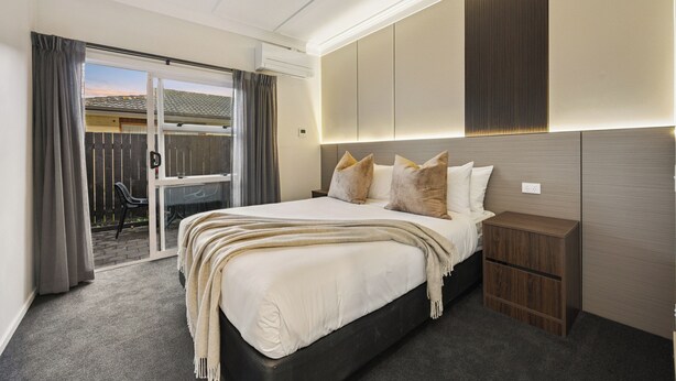 A bedroom with a large bed, a wooden nightstand, a sliding glass door leading to a patio, and a built-in headboard with integrated lighting. - Aroha Motel (Auckland)