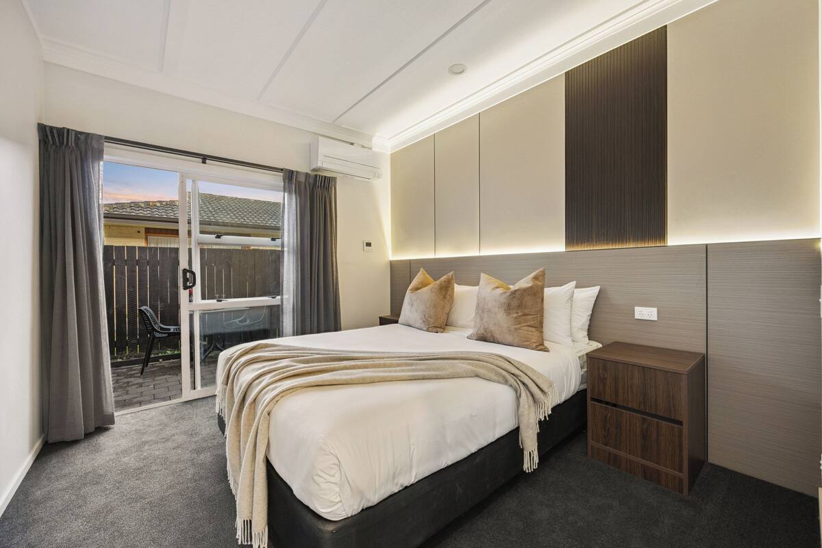 A bedroom with a large bed, a wooden nightstand, a sliding glass door leading to a patio, and a built-in headboard with integrated lighting.