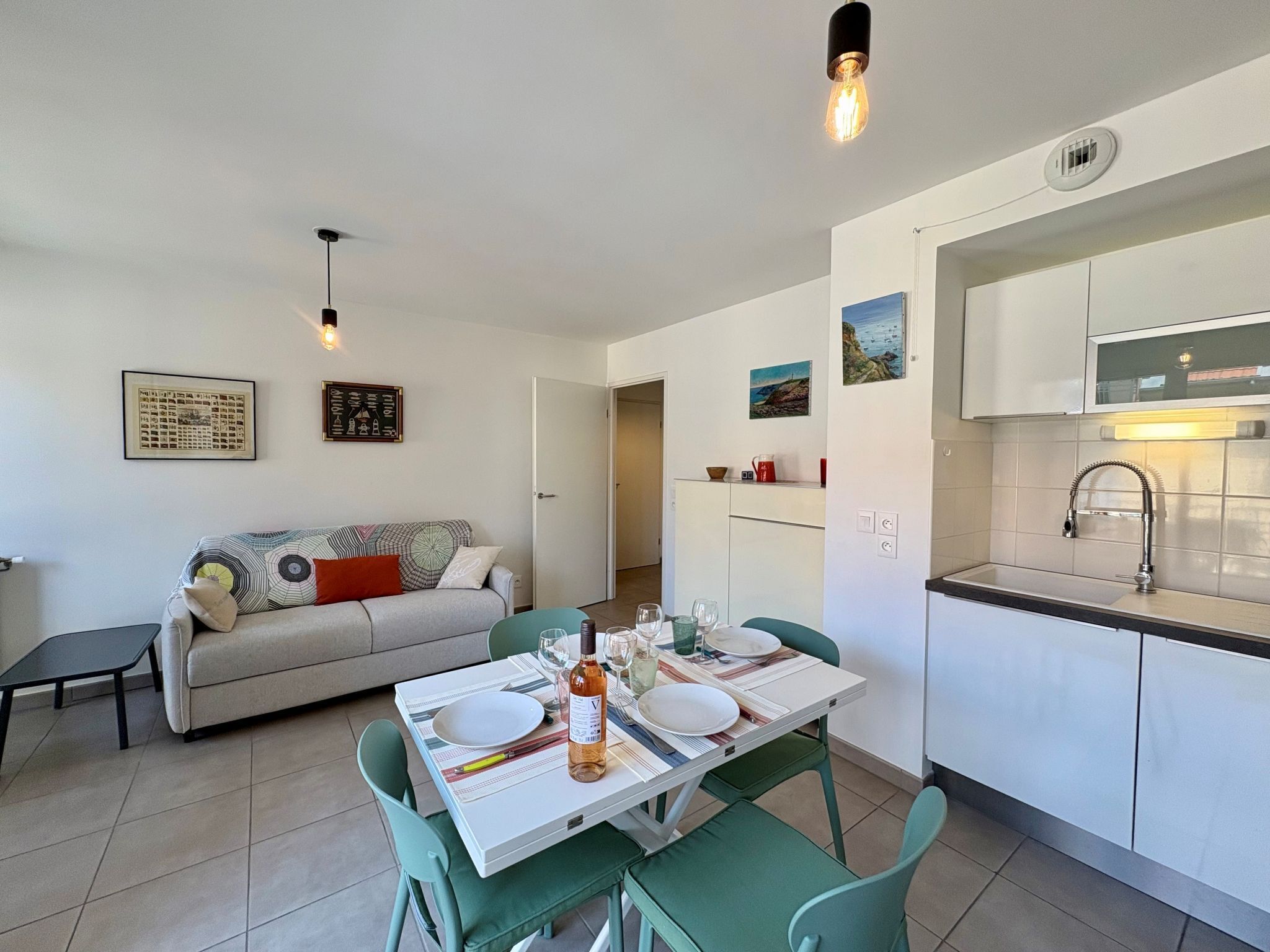 Une cuisine moderne avec des meubles blancs, un coin repas avec une table dressée pour quatre personnes, et un salon avec un canapé et des coussins.