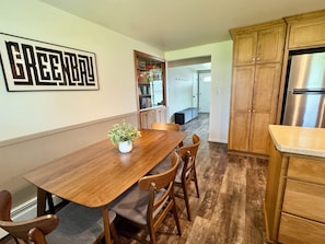 A dining area with a wooden table, chairs, a potted plant, and a kitchen with wooden cabinets and a stainless steel refrigerator.