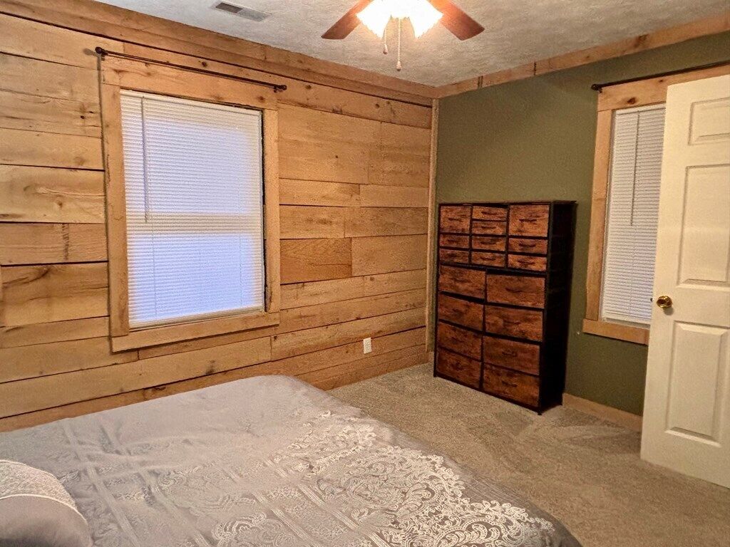 Una habitación con paredes de madera, una cama con colcha estampada, un aparador con cajones y un ventilador de techo.