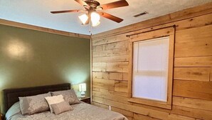 Ein Schlafzimmer mit Holzwand, einem Bett mit Kopfteil, einem Deckenventilator und einem Fenster mit Jalousien.