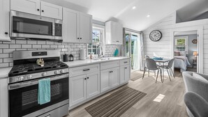 A modern kitchen with white cabinetry, a built-in microwave, a stove, and a dining area with a table and chairs. - Beachside Bliss at Sunset Retreat (West Dennis)