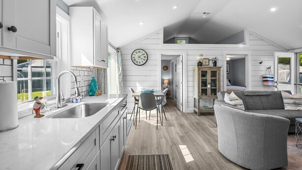 A modern kitchen with white cabinets, a stainless steel sink, and a dining area with a gray chair and table. - Beachside Bliss at Sunset Retreat (West Dennis)