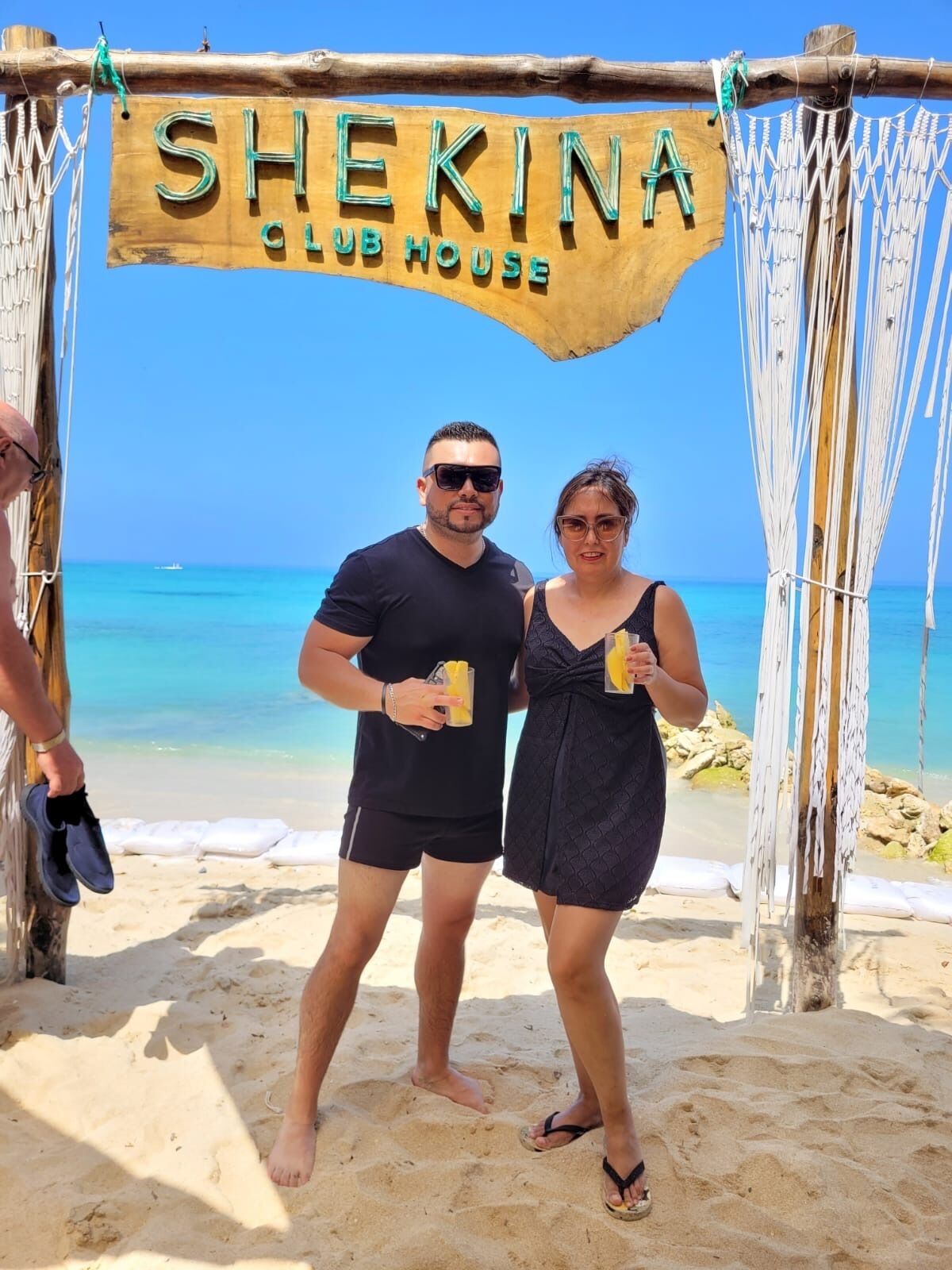 Une enseigne en bois sur laquelle on peut lire « Shekina Club House », placée sur fond de plage.