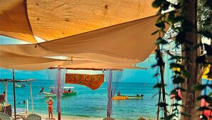 Un área de descanso frente a la playa con sillones y mesas, un letrero que dice 'She Kina Club House', y barcos en el agua.