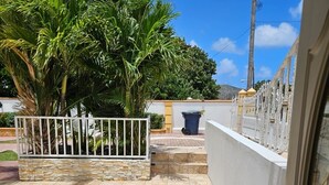 A patio with stone tiles, a white metal gate, and a view of a house with a yellow wall and a blue trash bin.