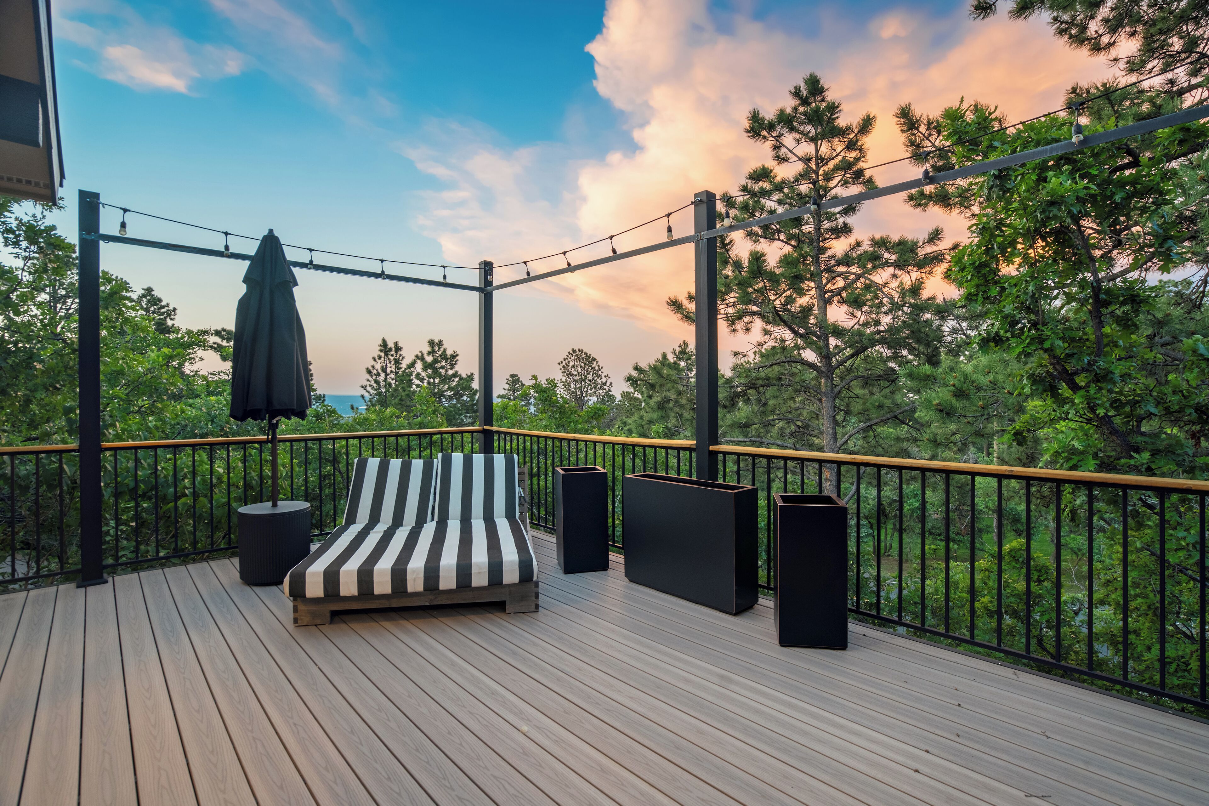 A wooden deck with a striped lounge chair, a hanging umbrella, and two black speakers.