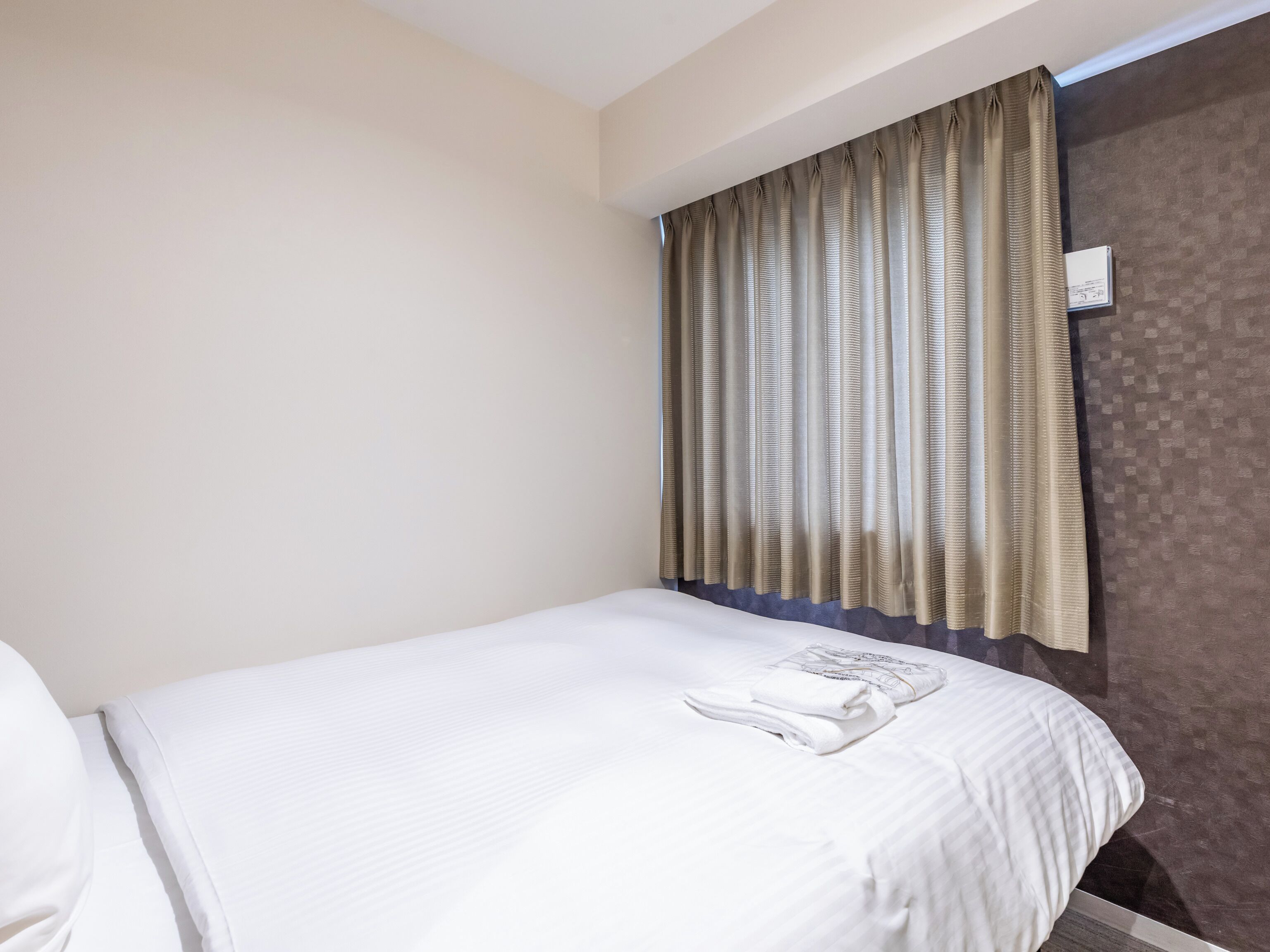 A hotel room with a single bed, a window with curtains, and a wall-mounted thermostat.