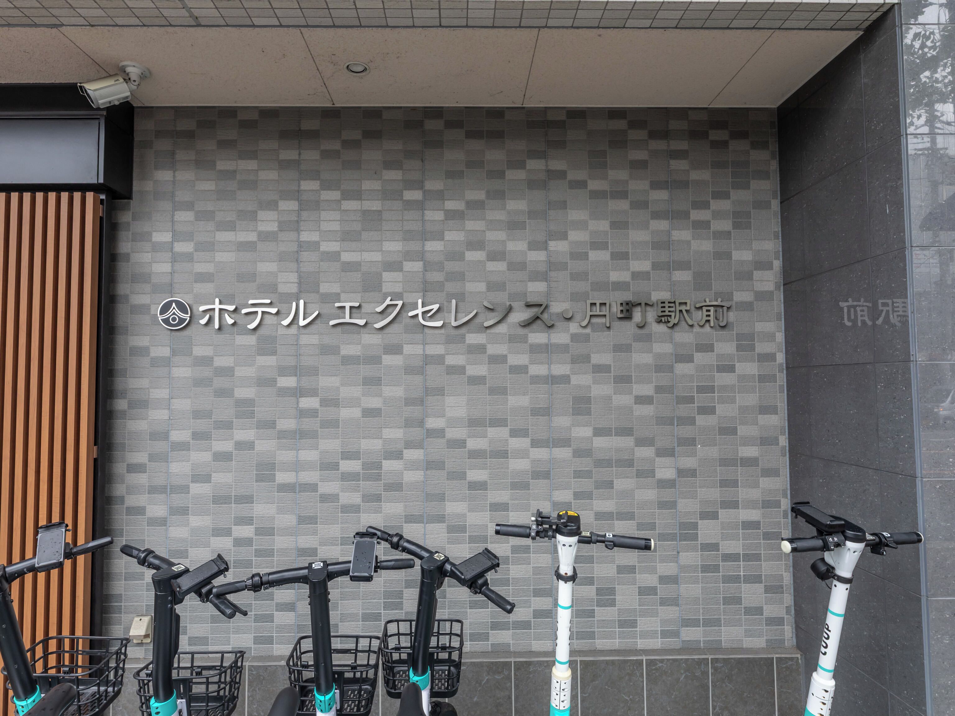 Una fila de patinetes eléctricos estacionados frente a un edificio con un muro de azulejos y un cartel en japonés.
