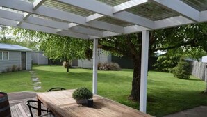 A covered patio area with a wooden table and chairs, a small potted plant, and a grassy yard with trees and a shed in the background. - Southern Comfort Twizel Escape (Twizel)
