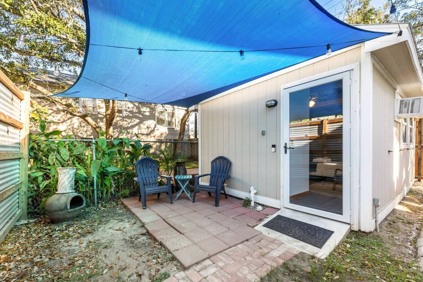Ein kleiner, abgeschlossener Patio-Bereich mit einem blauen Sonnensegel, zwei Adirondack-Stühlen und einer weißen Tür, die zu einer Garage führt.