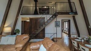 Ein großzügiger Wohnbereich mit hoher Decke, einer Treppe zu einer oberen Ebene, Ledersofas, einem Esstisch für vier Personen und einem großen Fenster, das natürliches Licht hereinlässt.