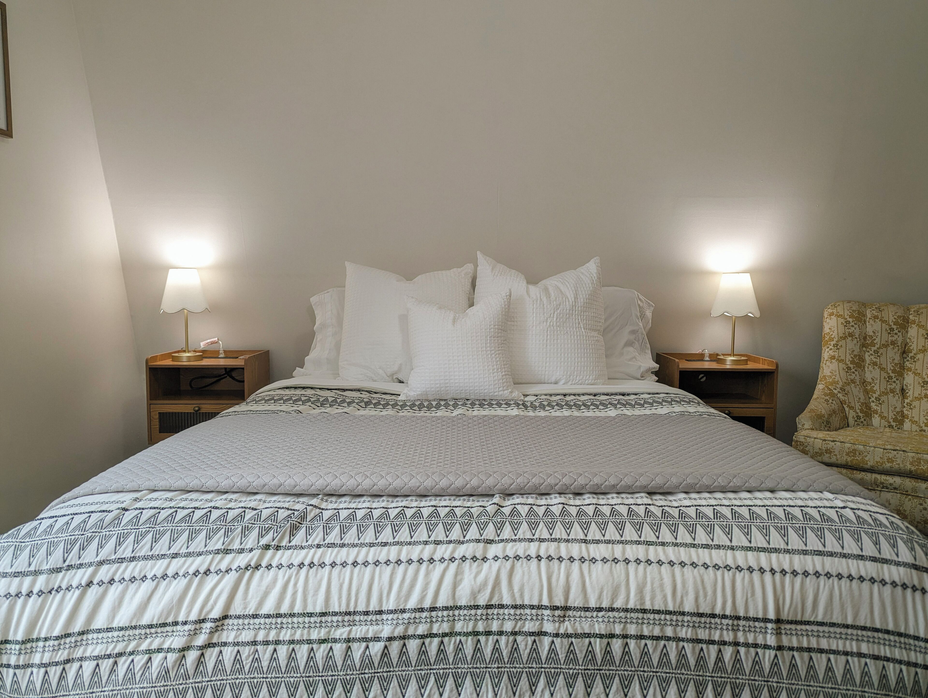 Ein Bett mit einem gemusterten Überwurf, zwei Nachttischlampen, ein Holznachttisch und ein floraler Sessel.