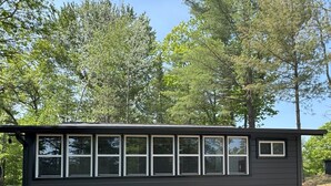 A small, dark-colored cabin with multiple windows, surrounded by trees and a grassy area. - Waubee Lake Remodeled Cozy Cottage on ATV Trails (Lakewood)
