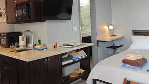 Ein kompaktes Hotelzimmer mit Küchenzeile, einem Bett mit weißer Tagesdecke, einem kleinen Tisch mit Lampe und einem Sessel.