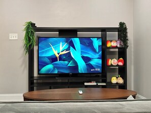 Ein modernes Wohnzimmer mit einem großen Flachbildfernseher an einer schwarzen Unterhaltungseinheit, einem Holztisch in der Mitte und einem Regal mit dekorativen Gegenständen rechts.