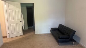 Une pièce avec une moquette beige, un canapé en cuir noir, une porte blanche et un couloir menant à une autre pièce.