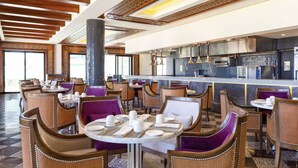 Ein geräumiges Restaurant mit runden Tischen, gepolsterten Stühlen, einer zentralen Säule und einer Küche im Hintergrund.
