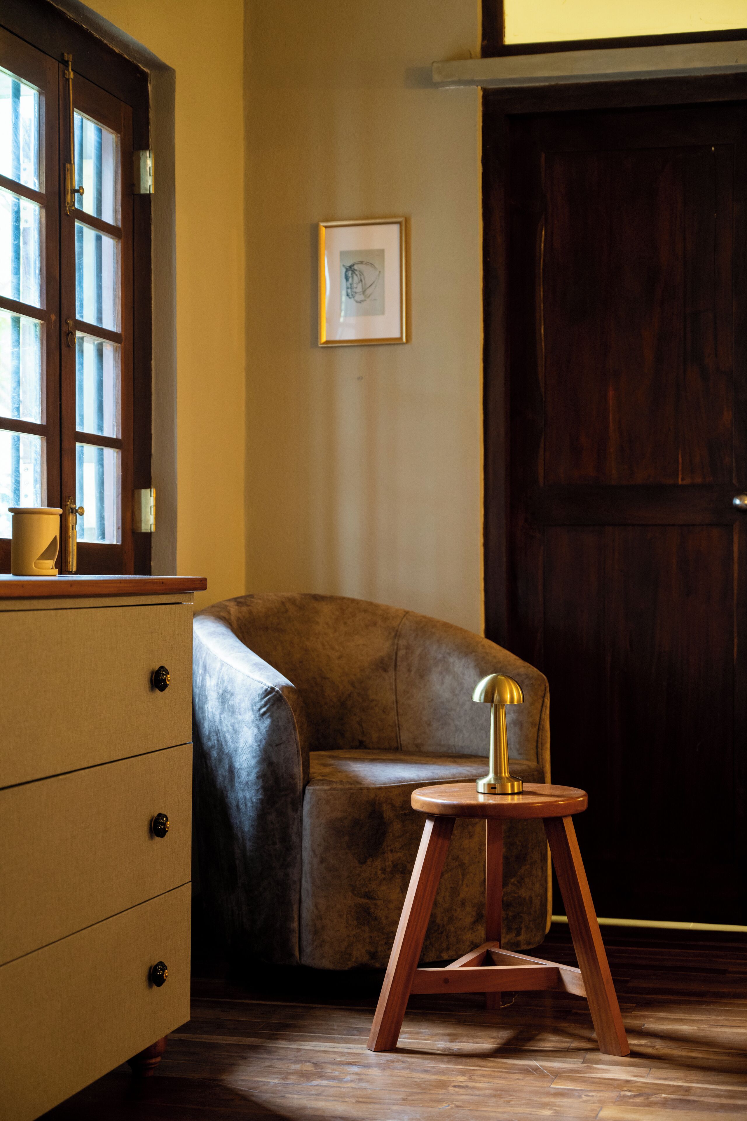 Una habitación con piso de madera, un sillón de cuero marrón, una mesita de madera con lámpara, un armario de madera y un cuadro enmarcado en la pared.