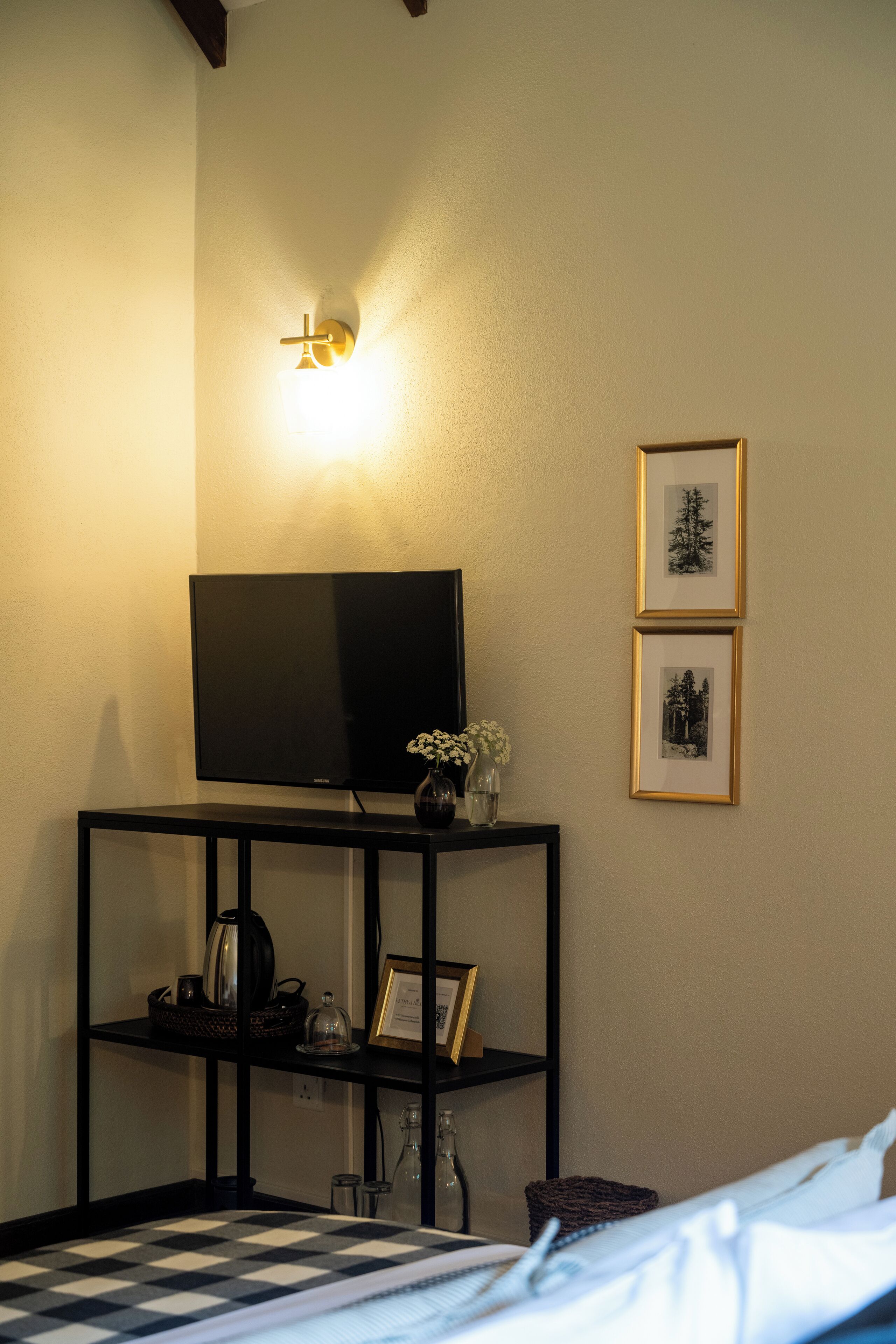 A bedroom with a bed, a TV on a stand, a small table with a vase and picture frames, and a wall-mounted light.