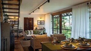 A cozy living room with a sofa, a dining table set for a meal, and a large window with curtains. - Tathya Hills - Luxe Garden Retreat (Nuwara Eliya)