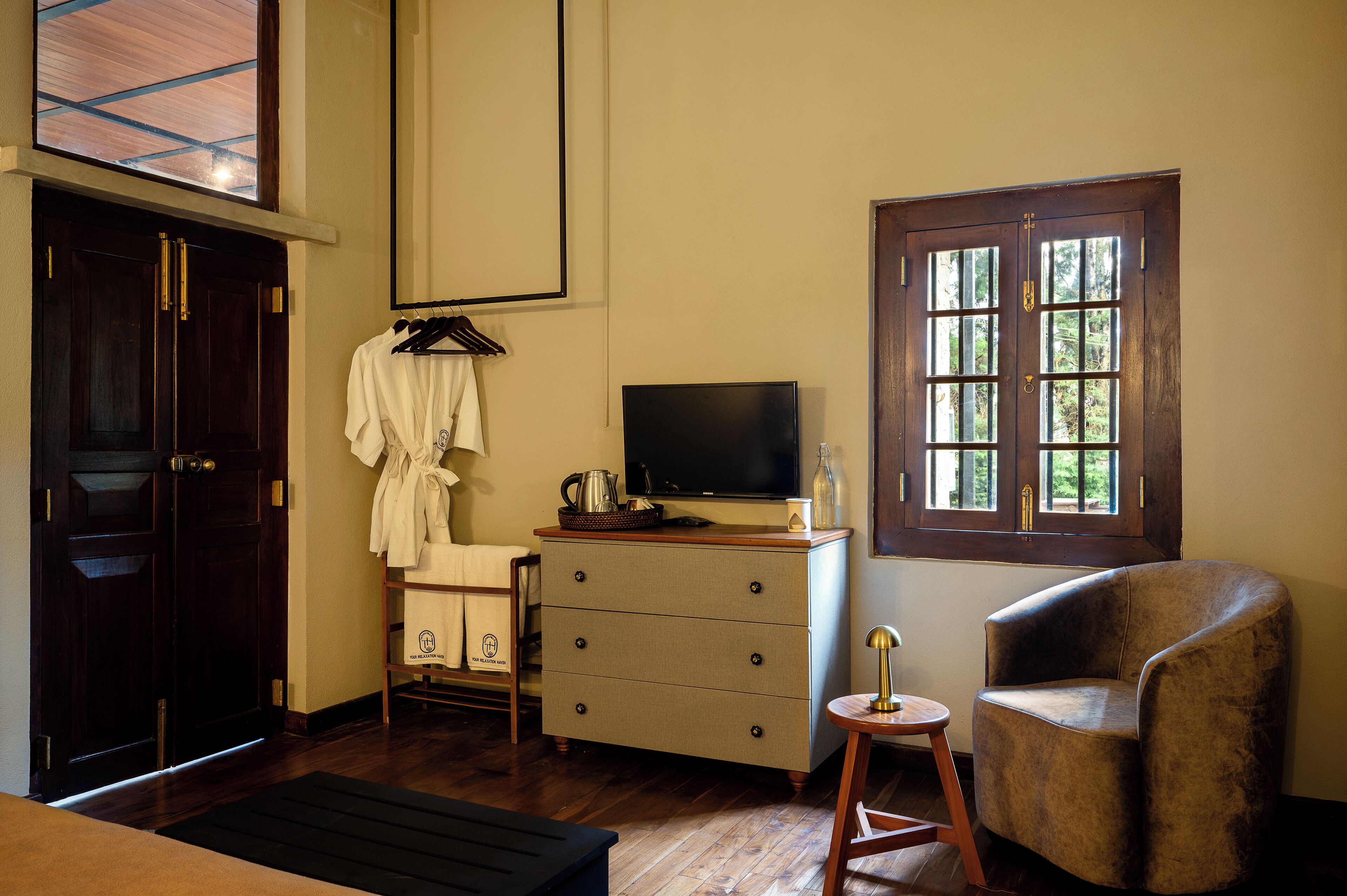 A hotel room with a wooden floor, a television on a dresser, a window with multiple panes, a chair, and a coat rack with a robe.